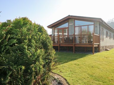 A small wooden house with a deck and glass windows surrounded by grass and bushes at Tranquillity - Heritage Park in Stepaside near Kilgetty