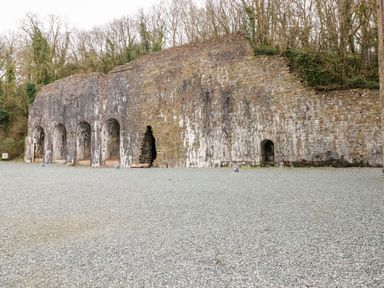 A stone wall with multiple arched openings and a gravel ground in front with trees behind at Tranquillity - Heritage Park in Stepaside near Kilgetty