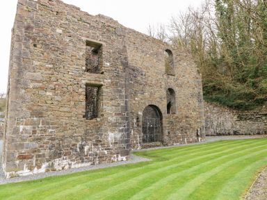 The exterior wall of an old stone building with arched and rectangular window openings next to a grassy lawn and trees at Tranquillity - Heritage Park in Stepaside near Kilgetty