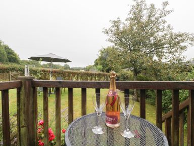 A garden with a round metal table holding a bottle and two glasses on a wooden deck at Floras Barn in Crowcombe