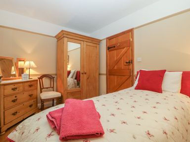 A bedroom with a bed with floral bedding red pillows wooden wardrobe dresser chair and wooden door at Floras Barn in Crowcombe