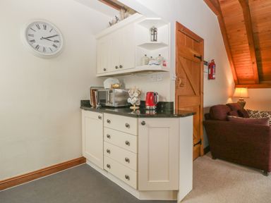 A small kitchen area with white cabinets a microwave a red kettle and a clock on the wall next to a living room with a maroon sofa and a wooden door at Floras Barn in Crowcombe