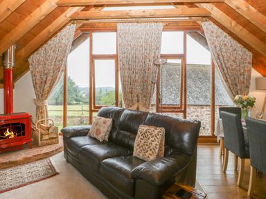 A living room with a black leather sofa a red wood-burning stove floral curtains and a dining table with chairs at Floras Barn in Crowcombe