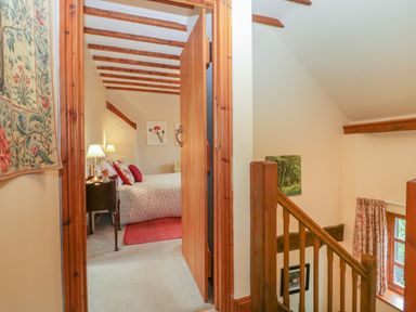 A bedroom with a bed and nightstand visible through a doorway next to a staircase with wooden railing at Floras Barn in Crowcombe