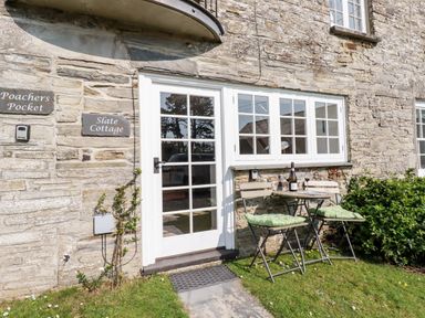 A stone cottage exterior with a white framed glass door and windows a small table with two chairs and cushions on grass at Slate Cottage Tredethy near Bodmin Moor