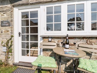 A small outdoor table with two chairs and a bottle of prosecco outside a stone cottage with white framed door and windows at Slate Cottage Tredethy near Bodmin Moor