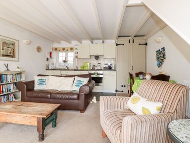 A living room with a brown leather sofa, striped armchair, wooden coffee table, bookshelf, and a kitchen area at Slate Cottage in Tredethy near Bodmin Moor