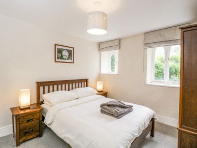 A bedroom with a wooden bed frame and white bedding two bedside tables with lamps a wooden wardrobe and two windows at Viaduct House in Lydbrook