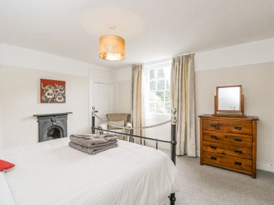 A bedroom with a metal bed frame and white bedding a wooden dresser with a mirror an armchair and a window with curtains at Viaduct House in Lydbrook