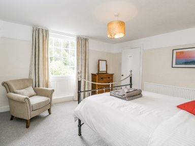 A bedroom with a metal bed frame, an armchair, a wooden dresser with a mirror, curtains, and a framed picture on the wall at Viaduct House in Lydbrook