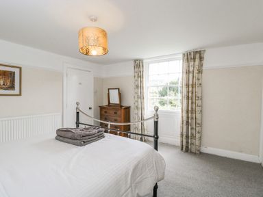 A bedroom with a metal bed frame a white bedspread folded towels on the bed a wooden chest of drawers with a small mirror a window with patterned curtains and a ceiling light at Viaduct House in Lydbrook