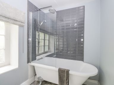 A bathroom with a clawfoot bathtub shower combination glass shower screen grey tiled wall and a window with a Roman blind at Viaduct House in Lydbrook