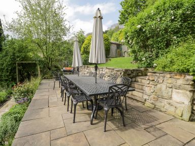 An outdoor stone patio with a long black metal table and chairs two closed umbrellas and a garden shed at Viaduct House in Lydbrook