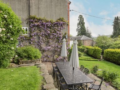An outdoor patio with a long black metal table and chairs under closed umbrellas next to a garden with purple flowering vines on a wall at Viaduct House in Lydbrook
