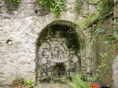 A small outdoor seating area with a round table and two metal chairs against a stone wall with potted plants and greenery at Viaduct House in Lydbrook