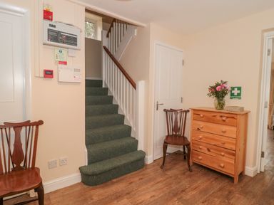 An entry hallway with stairs carpeted in green a wooden chest of drawers with flowers on top and wooden chairs at Aldrich Cottage in Radstock