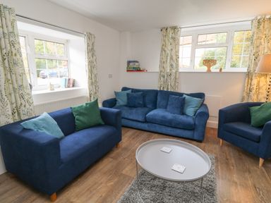 A living room with blue sofas green cushions a round gray table and windows with patterned curtains at Aldrich Cottage in Radstock
