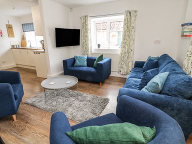 A living room with blue sofas and a round gray table on a rug with a kitchen area in the background at Aldrich Cottage in Radstock
