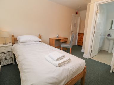 A bedroom with a single bed with white bedding a bedside table with a lamp a wooden desk and chair a chest of drawers and an open door leading to a bathroom at Aldrich Cottage in Radstock