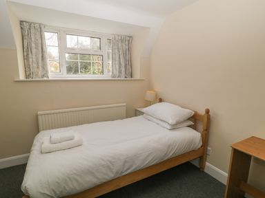 A small bedroom with a single wooden bed with white bedding next to a window with curtains a bedside table with a lamp and a wooden desk at Aldrich Cottage in Radstock