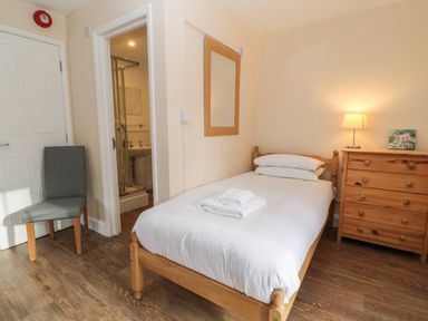 A bedroom with a single bed and white bedding a wooden chest of drawers with a lamp and a chair near a door at Aldrich Cottage Radstock