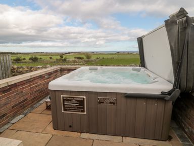 An outdoor hot tub with bubbling water on a stone patio with a view of green fields and the sea at West Gate in Embleton