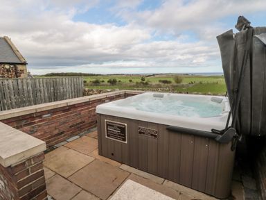 An outdoor hot tub on a stone patio with a wooden fence and grassy fields in the background at West Gate in Embleton