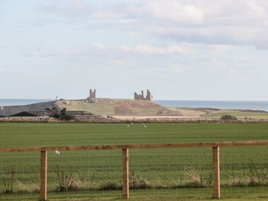 A green field with a wooden fence in the foreground and castle ruins near the sea in the background at West Gate in Embleton