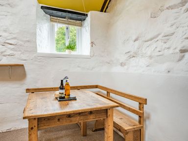 A corner with a wooden table and bench under a window with a plant on the sill at Celyn Farm Cottage in Deiniolen near Llanberis