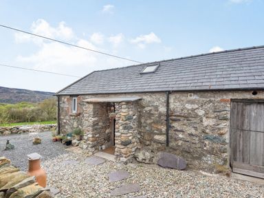A stone cottage with a slate roof and a gravel courtyard with paving stones and potted plants at Celyn Farm Cottage in Deiniolen near Llanberis