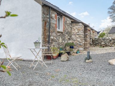 An outdoor gravel area with a small table and two chairs next to a stone and white plaster building with potted plants at Celyn Farm Cottage in Deiniolen near Llanberis