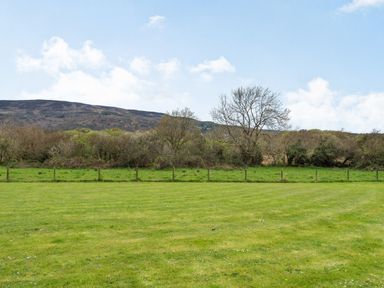 A grassy field with a wooden fence and trees in front of hills under a blue sky at Celyn Farm Cottage in Deiniolen near Llanberis