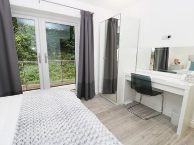 A bedroom with a bed covered by a textured blanket, gray curtains, a mirrored wardrobe, a white desk with a mirror, and a transparent chair at The Office Gardens Apartment 2 Whitby