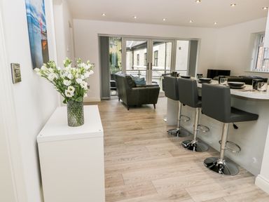 An open-plan living area with a white cabinet holding a vase of white flowers a kitchen island with black bar stools and a dining setup and a seating area with chairs by glass doors at The Office Gardens Apartment 2 Whitby