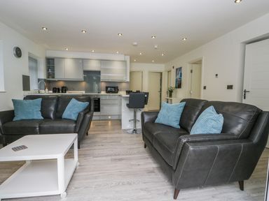 A living room with two black leather sofas and blue pillows a white coffee table and kitchen area in the background at The Office Gardens Apartment 2 in Whitby