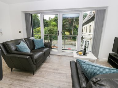 A living room with black leather sofas and blue cushions facing glass doors overlooking greenery at The Office Gardens Apartment 2 in Whitby