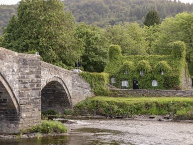 A stone bridge over a river with a house covered in ivy and trees in the background at Dale Cottage in Llanrwst