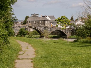 A stone arch bridge over a river with green grass and a paved path leading to it at Dale Cottage in Llanrwst
