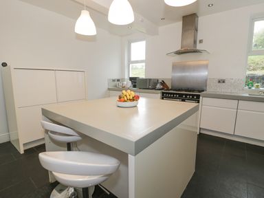 A kitchen with a central island with fruit bowl two bar stools white cabinets and stainless steel stove at Dale Cottage in Llanrwst