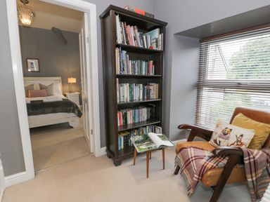 A small reading nook with a brown armchair and bookshelf next to a window and a view into a bedroom at Dale Cottage in Llanrwst