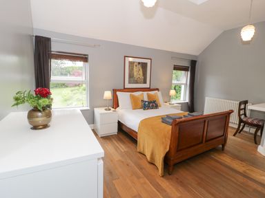 A bedroom with a wooden bed with yellow bedding two bedside tables with lamps a white dresser with a vase with flowers and two windows at Dale Cottage in Llanrwst
