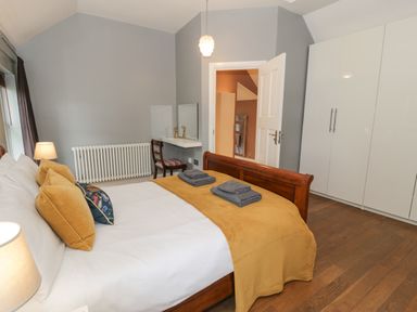 A bedroom with a wooden bed with yellow and floral pillows a yellow blanket two folded towels a white wardrobe a chair a desk with a mirror and a lamp at Dale Cottage in Llanrwst