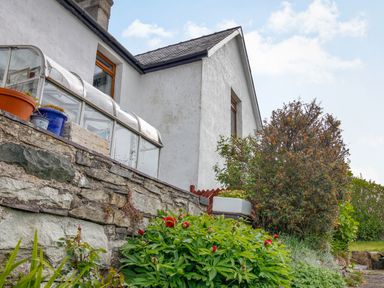 The exterior of a white house with a stone wall and various plants and bushes at Pen Y Graig in Talysarn