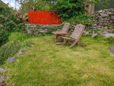 A garden area with two wooden chairs and a small wooden table on grass with stone walls and a red fence in the background at Pen Y Graig in Talysarn