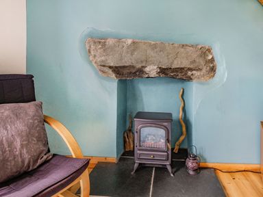 A small stove inside a stone fireplace with a wooden chair and cushion nearby at Pen Y Graig in Talysarn