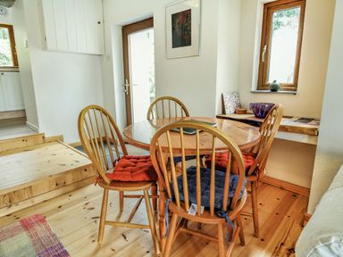 A dining area with a round wooden table surrounded by four wooden chairs with cushions near windows and a door at Pen Y Graig in Talysarn