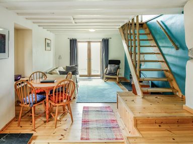 A dining area with a wooden table and four chairs and a living room with a sofa and armchair next to a wooden staircase at Pen Y Graig in Talysarn