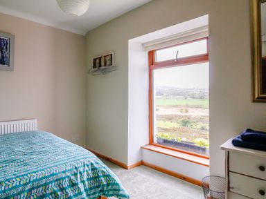 A bedroom with a patterned blue bedspread a window with a wooden frame showing a rural landscape and a white dresser with folded blue towels at Pen Y Graig in Talysarn