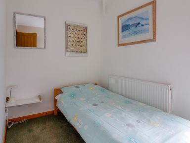 A small bedroom with a single bed with blue floral bedding a wall mirror a framed picture a lamp on a floating white shelf and a radiator at Pen Y Graig in Talysarn