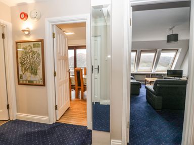 A hallway with blue carpet and open doors leading to a dining area with dining table and chairs and a living room with sofas and windows at Rothay 17 in Ambleside